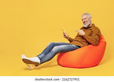 Full Size Body Length Fun Elderly Gray-haired Bearded Man 60s Years Old Wear Brown Shirt Sit In Bag Chair Hold Use Point Finger On Mobile Cell Phone Isolated On Plain Yellow Background Studio Portrait