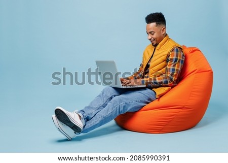 Full size body length fun young black man 20s years old wears yellow waistcoat shirt sit in bag chair hold use work on laptop pc computer isolated on plain pastel light blue background studio portrait