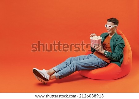 Full size body length fun young brunet man 20s wears red t-shirt green jacket sit in bag chairin 3d glasses watch movie film hold bucket of popcorn isolated on plain orange background studio portrait.