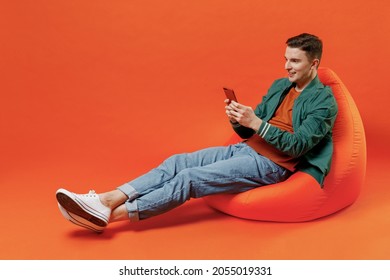 Full size body length happy smiling vivid young brunet man 20s wears red t-shirt green jacket sit in bag chair hold in hand use mobile cell phone isolated on plain orange background studio portrait.