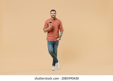 Full Size Body Length Happy Tattooed Young Brunet Man 20s Wear Apricot Shirt Stand Hold Takeaway Delivery Craft Paper Brown Cup Coffee To Go Isolated On Light Pastel Orange Background Studio Portrait