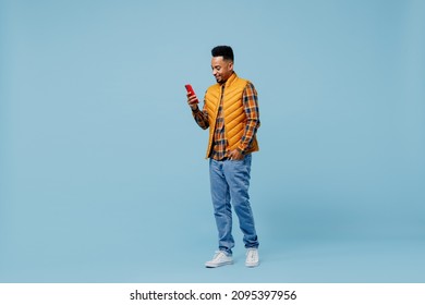 Full size body length happy young black man 20s wears yellow waistcoat shirt hold use mobile cell phone typing browsing chatting send sms isolated on plain pastel light blue background studio portrait