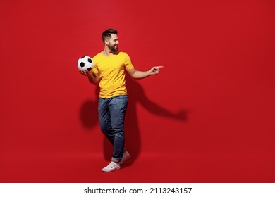 Full Size Body Length Happy Excited Young Bearded Man Football Fan In Yellow T-shirt Hold Soccer Ball Point On Workspace Area Copy Space Mock Up Isolated On Plain Dark Red Background Studio Portrait