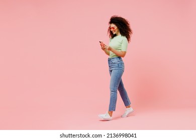 Full Size Body Length Happy Fancy Young Curly Latin Woman 20s Wears Casual Clothes Sunglasses Go Move Hold In Hand Use Mobile Cell Phone Isolated On Plain Pastel Light Pink Background Studio Portrait