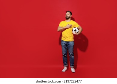 Full size body length happy young bearded man football fan in yellow t-shirt support favorite team hold soccer ball sing national country anthem isolated on plain dark red background studio portrait
