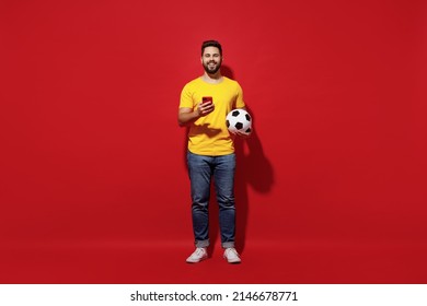 Full Size Body Length Happy Young Bearded Man Football Fan In Yellow T-shirt Cheer Up Support Favorite Team Hold Soccer Ball Use Mobile Cell Phone Isolated On Plain Dark Red Background Studio Portrait