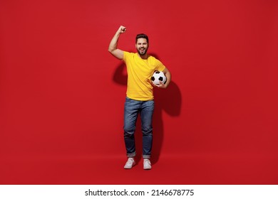 Full Size Body Length Happy Young Bearded Man Football Fan In Yellow T-shirt Cheer Up Support Favorite Team Hold Soccer Ball Doing Winner Gesture Isolated On Plain Dark Red Background Studio Portrait