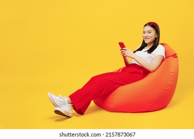 Full Size Body Length Happy Young Woman Of Asian Ethnicity 20s Years Old Wears Casual Clothes Sit In Bag Chair Hold Use Mobile Cell Phone Rest Isolated On Plain Yellow Background Studio Portrait