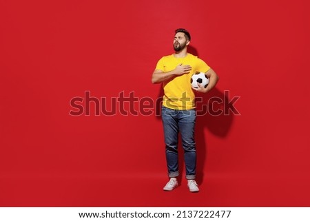 Full size body length happy young bearded man football fan in yellow t-shirt support favorite team hold soccer ball sing national country anthem isolated on plain dark red background studio portrait
