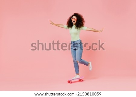Full size body length happy fun excited bright young curly latin woman 20s wears casual clothes sunglasses skate on board spreading hands isolated on plain pastel light pink background studio portrait
