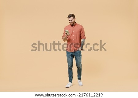Full size body length happy young man 20s he wears orange shirt go stride walk hold in hand use mobile cell phone isolated on plain pastel light beige background studio portrait. Tattoo translate fun