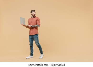 Full Size Body Length Joyful Tattooed Young Brunet Man 20s Short Haircut Wears Apricot Shirt Go Stride Hold Use Laptop Pc Computer Look Back Behind Isolated On Pastel Orange Background Studio Portrait