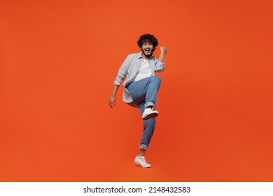 Full size body length jubilant exultant young bearded Indian man 20s years old wears blue shirt do winner gesture celebrate clenching fists say yes isolated on plain orange background studio portrait