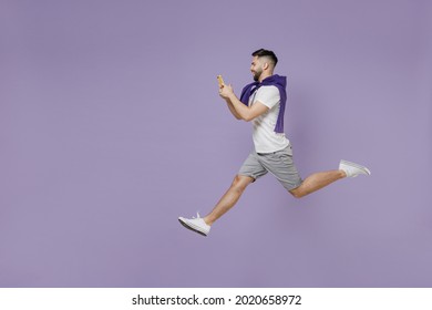 Full size body length side view profile satisfied young brunet man 20s wears white t-shirt purple shirt jump hold in hand use mobile cell phone isolated on pastel violet background studio portrait.