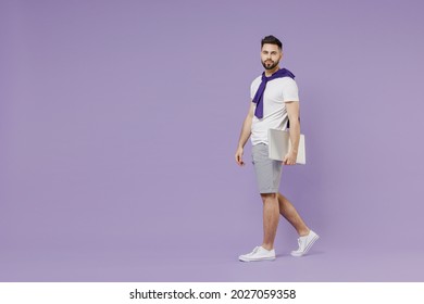 Full size body length side view serious young brunet man 20s wears white t-shirt purple shirt hold under hand laptop pc computer go move stroll isolated on pastel violet background studio portrait.