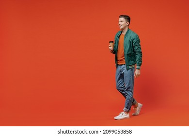 Full size body length side view young brunet man 20s wears red t-shirt green jacket go step hold takeaway delivery craft paper brown cup coffee to go isolated plain orange background studio portrait.