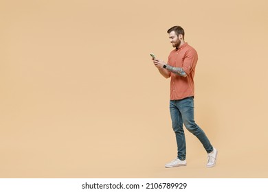 Full size body length side view happy tatooed young brunet man 20s short haircut wears apricot shirt go stride hold in hand use mobile cell phone isolated on pastel orange background studio portrait. - Shutterstock ID 2106789929