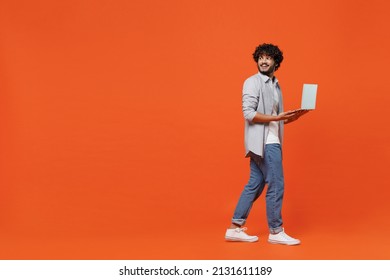 Full size body length side view young bearded Indian man 20s wears blue shirt hold use work on laptop pc computer typing browsing chatting send sms isolated on plain orange background studio portrait - Shutterstock ID 2131611189