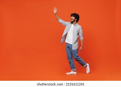 Full size body length side profile view cheerful young bearded Indian man 20s years old wears blue shirt meet greet waving hand as notices someone isolated on plain orange background studio portrait