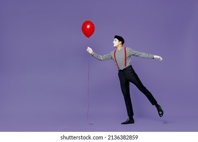 Full size body length side view young mime man with white face mask wears striped shirt beret hold colorful air inflated helium balloon isolated on plain pastel light violet background studio portrait