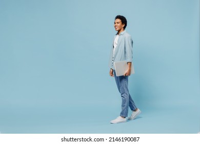 Full size body length side view young black curly man 20s wears white shirt hold use work on laptop pc computer stepping strolling pace isolated on plain pastel light blue background studio portrait