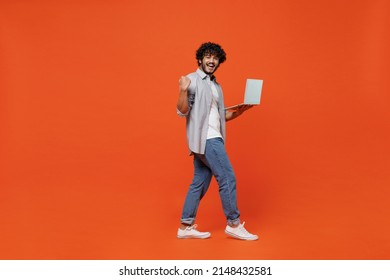 Full size body length side view young bearded Indian man 20s wears blue shirt hold use work on laptop pc computer do winner gesture clenching fists isolated on plain orange background studio portrait