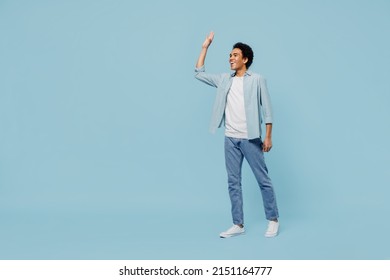 Full size body length side view young black curly man 20s years old wears white shirt meet greet waving hand as notices someone strolling isolated on plain pastel light blue background studio portrait