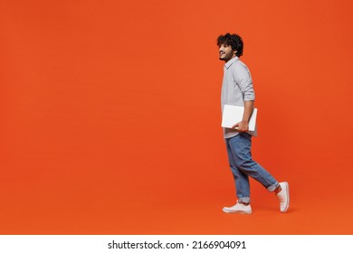 Full size body length side profile view young bearded Indian man 20s years old wears blue shirt hold use work on laptop pc computer strolling pace isolated on plain orange background studio portrait