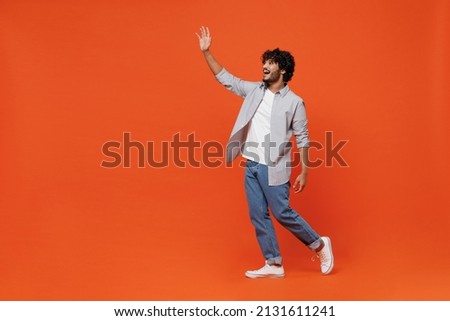 Full size body length side profile view cheerful young bearded Indian man 20s years old wears blue shirt meet greet waving hand as notices someone isolated on plain orange background studio portrait