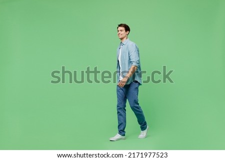 Full size body length side view blithesome charismatic stunning delight young brunet man 20s years old wears blue shirt looking aside strolling pace isolated on plain green background studio portrait
