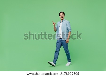 Full size body length side view fun young brunet man 20s years old wears blue shirt pointing index finger aside on workspace area copy space mock up isolated on plain green background studio portrait