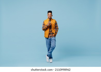 Full size body length smiling young black man 20s wears yellow waistcoat shirt hold takeaway delivery craft paper brown cup coffee to go isolated on plain pastel light blue background studio portrait