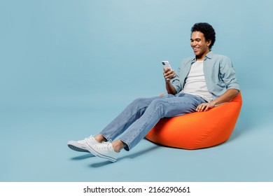 Full size body length smiling young black curly man 20s years old wears white shirt sit in bag chair hold in hand use mobile cell phone isolated on plain pastel light blue background studio portrait - Shutterstock ID 2166290661
