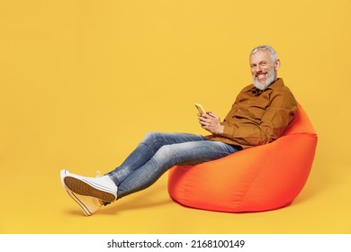 Full Size Body Length Smiling Elderly Gray-haired Bearded Man 50s Years Old He Wears Brown Shirt Sit In Bag Chair Hold In Hand Use Mobile Cell Phone Isolated On Plain Yellow Background Studio Portrait