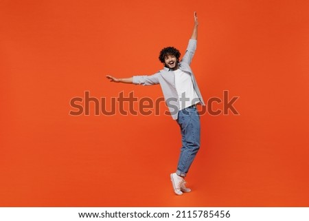 Full size body length smiling young bearded Indian man 20s years old wears blue shirt standing on toes dancing lean back have fun spreading hands isolated on plain orange background studio portrait