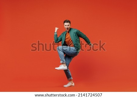 Full size body length smiling vivid fun young brunet man 20s wears red t-shirt green jacket doing winner gesture celebrating clenching fists say yes isolated on plain orange background studio portrait