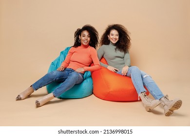 Full Size Body Length Two Smiling Charming Fun Young Curly Black Women Friends 20s Wear Casual Shirts Clothes Sit In Bag Chair Looking Camera Isolated On Plain Pastel Beige Background Studio Portrait