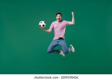 Full size body length young fun man fan wear pink t-shirt cheer up support football sport team hold in hand soccer ball watch tv live stream scream jump isolated on dark green color background studio