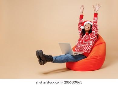 Full size body length young african american Santa woman in Christmas hat sweater sit in bag chair hold use work on laptop pc computer stretching isolated on pastel beige background studio portrait - Powered by Shutterstock