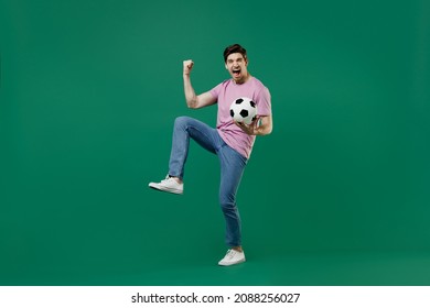 Full size body length young fun man fan wear basic pink t-shirt cheer up support football sport team hold in hand soccer ball watch tv live stream scream isolated on dark green color background studio