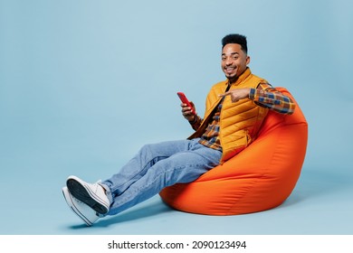 Full Size Body Length Young Black Man 20s Years Old Wears Yellow Waistcoat Shirt Sit In Bag Chair Hold Use Pointing On Mobile Cell Phone Isolated On Plain Pastel Light Blue Background Studio Portrait