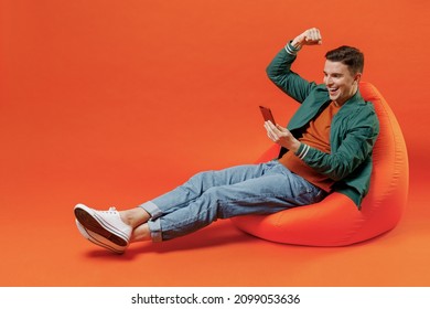 Full size body length young brunet man 20s wears red t-shirt green jacket sit in bag chair hold in hand use mobile cell phone celebrate clench fists isolated on plain orange background studio portrait - Powered by Shutterstock