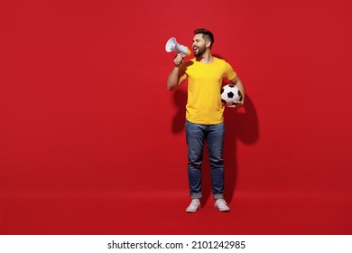 Full size body length young bearded man football fan in yellow t-shirt cheer up support favorite team hold soccer ball scream aside in megaphone isolated on plain dark red background studio portrait