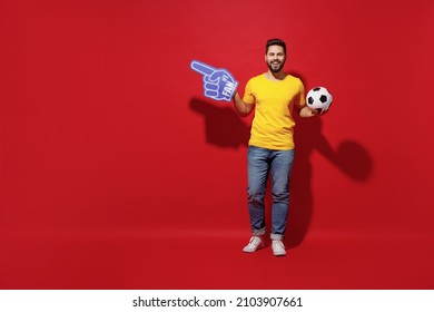 Full Size Body Length Young Man Football Fan In Yellow T-shirt Hold Soccer Ball Point Fan Foam Glove Finger On Workspace Area Copy Space Mock Up Isolated On Plain Dark Red Background Studio Portrait