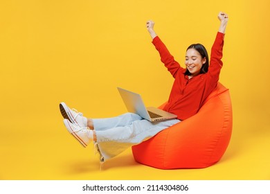 Vollgroße Körperlänge junge Frau von asiatischen ethnischen 20ern in ungezwungenen Kleidung sitzen in Sackstuhl verwenden Arbeit auf Laptop PC-Computer mit Gewinner-Geste einzeln auf einfarbgelbegelbem Hintergrund Studioportrait