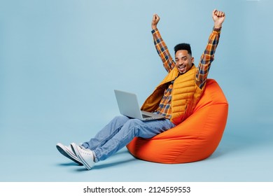 Full size body length young black man 20s wears yellow waistcoat shirt sit in bag chair hold laptop pc computer just found out great win isolated on plain pastel light blue background studio portrait