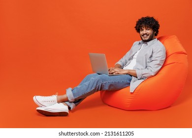Full size body length young bearded Indian man 20s years old wears blue shirt sit in bag chair hold use work on laptop pc computer chatting send sms isolated on plain orange background studio portrait
