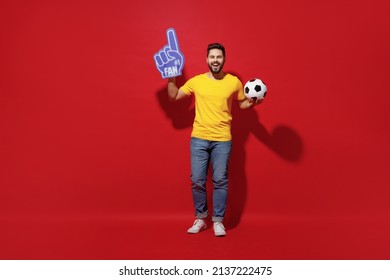 Full size body length young bearded man football fan in yellow t-shirt cheer up support favorite team hold soccer ball fan foam glove finger up isolated on plain dark red background studio portrait
