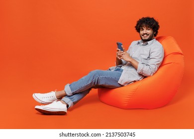 Full Size Body Length Young Bearded Indian Man 20s Years Old Wears Blue Shirt Sit In Bag Chair Hold In Hand Use Mobile Cell Phone Chatting Send Sms Isolated On Plain Orange Background Studio Portrait