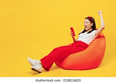 Full size body length young girl woman of Asian ethnicity 20s years old in casual clothes sit in bag chair hold use mobile cell phone do winner gesture isolated plain yellow background studio portrait - Shutterstock ID 2138216025